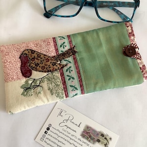 May include: Blue tortoise shell reading glasses with clear lenses and a rectangular fabric eyeglass case featuring a patchwork design of a robin bird, floral patterns, and green fabric, secured with a brown button and a business card.