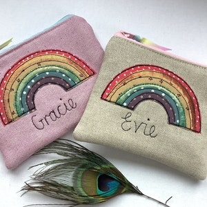 Può includere: Due piccole pochette con cerniera con ricamo a forma di arcobaleno. La pochette a sinistra è rosa con il nome "Gracie" ricamato sotto l'arcobaleno. La pochette a destra è beige con il nome "Evie" ricamato sotto l'arcobaleno.