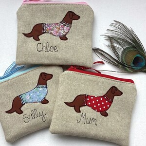 Könnte beinhalten: Drei kleine Reißverschlusstaschen aus beigefarbenem Leinenstoff mit gestickten Dackeln, die Pullover tragen. Die Taschen sind mit den Namen "Chloe", "Sally" und "Mum" personalisiert.