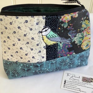 May include: A zippered pouch featuring a patchwork design with cream fabric with blue floral patterns, a black strip with a blue tit bird appliqué, and a dark floral panel. The bottom section is teal with a white dandelion seed head print.