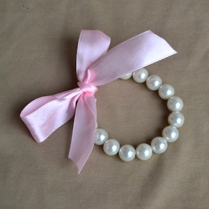 May include: A bracelet made of white, round pearls, tied with a light pink satin ribbon bow. The bow is on the left side of the bracelet, and the pearls are evenly spaced around the wrist. The background is a solid tan color.