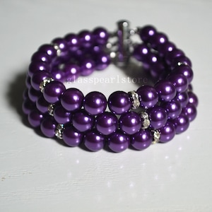Puede incluir: Una pulsera de tres hilos con cuentas brillantes de color púrpura intenso. La pulsera tiene un cierre plateado y está adornada con pequeños espaciadores de pedrería brillantes. El texto "glasspearlstore" es visible en la pulsera.