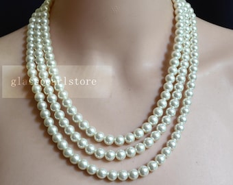 May include: Three-strand ivory pearl necklace. The necklace features evenly spaced, round pearls. The strands are draped around a neck, showcasing the necklace's design. The text "glasspearlstore" is visible on the left side of the image.