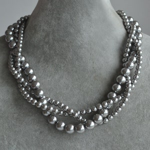 May include: A multi-strand necklace with graduated, silvery-grey beads. The beads vary in size, creating a layered effect. The necklace is displayed on a grey mannequin bust, showcasing its design and form.