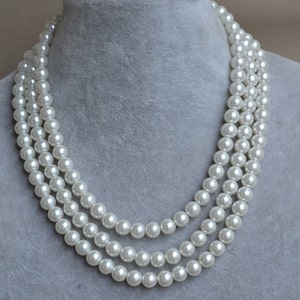 May include: A three-strand pearl necklace with white faux pearls. The necklace is displayed on a grey velvet background.
