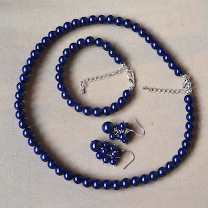 Puede incluir: Un conjunto de joyería compuesto por un collar y una pulsera de perlas sintéticas redondas de color azul intenso, junto con un par de pendientes de racimo a juego. El collar y la pulsera incluyen cierres de mosquetón y cadenas de extensión plateadas.