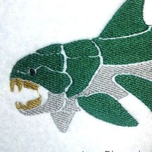 May include: Embroidered design of a green and silver prehistoric fish with a large mouth and sharp teeth. The design is stitched on a white fabric background.