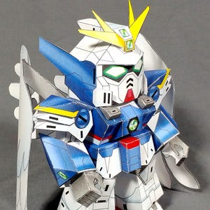 May include: A papercraft Gundam robot figure, featuring a white head with yellow accents, blue and white armor, and red feet. The figure has wings and is posed against a gray background. The text "FOLDATICK.COM" is visible.