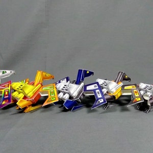 May include: Five colourful paper model spaceships, each with a different colour scheme. The spaceships are made of paper and have a futuristic design. They are arranged in a row, with the green and rainbow spaceship on the left and the red spaceship on the right.