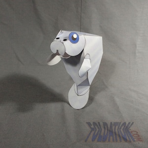 Puede incluir: Un manatí de papel colgante con un diseño geométrico. El manatí es principalmente blanco con acentos azules para los ojos. La manualidad de papel está suspendida por un hilo negro fino sobre un fondo gris.