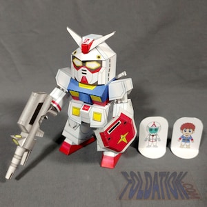 Puede incluir: Una figura de robot Gundam de papel blanco, rojo, azul y amarillo con una pistola plateada y un escudo rojo. También hay dos pequeños recortes de personajes y un logotipo. El robot mide aproximadamente 13 cm de altura.