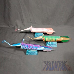 May include: Three colorful paper squid models. One is pink with orange accents, another is green and yellow, and the third is blue and purple. Each rests on a small blue stand with the text "Billy Monster".