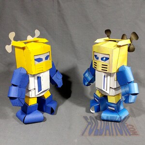 May include: Two papercraft robot figures. Each robot has a yellow head, blue arms and legs, and a white torso. The robots have a stylized design with angular shapes and details. One robot has white antlers, and the other has black antlers.