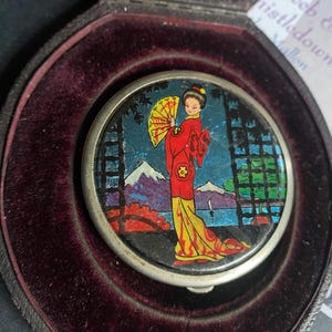 May include: A round silver-toned compact mirror featuring a colorful illustration of a person in a red and yellow kimono holding a yellow fan. The scene includes a mountain, water, and black lattice structures, all set within a dark velvet-lined case.