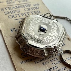 May include: An antique silver compact with intricate floral and geometric engravings. The compact features a small blue and white flag emblem with the letter "M". The background includes a vintage paper with text from "JOSEPH LEVY STEAMSHIP and SHIPS CREW OUTFITTER".
