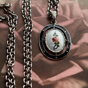 Könnte beinhalten: Silberfarbene Halskette mit ovalem Anhänger. Der Anhänger zeigt eine detaillierte Miniaturmalerei einer Rose. Der Anhänger ist von einem schwarzen Rand mit goldenen Akzenten umgeben. Die Kette ist im klassischen Kettenglied-Stil.