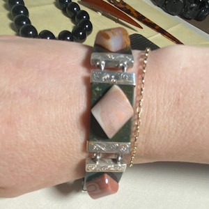 Op de afbeelding: Een armband met rechthoekige en ruitvormige edelstenen in bruin, roze en groen, verbonden door zilverkleurige metalen schakels. Een fijne gouden ketting sluit de armband.
