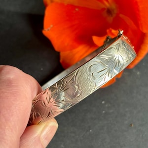 Puede incluir: Un brazalete rígido articulado de tono plateado con un patrón floral grabado en su superficie exterior. La banda de metal tiene un acabado pulido con líneas grabadas detalladas que forman motivos de hojas y pétalos, y un pequeño mecanismo de bisagra.