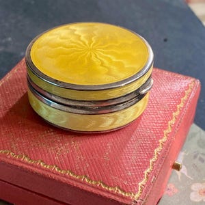 May include: A round, yellow enamel pill box with a silver-toned metal rim and a textured, sunburst-patterned lid. The small, circular container features a hinged design with a visible metal clasp on the side, showcasing a bright yellow finish.