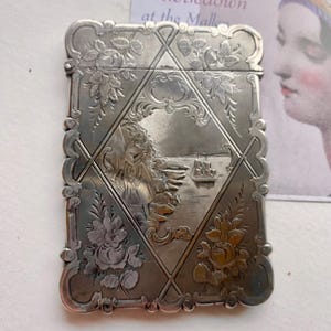 Könnte beinhalten: Ein rechteckiges silbernes Etui mit aufwendigen floralen und geometrischen Mustern. Die Vorderseite zeigt eine detaillierte Gravur einer Frau in der Nähe eines Gewässers mit einem Schiff. Das Etui ist wahrscheinlich Vintage oder antik.