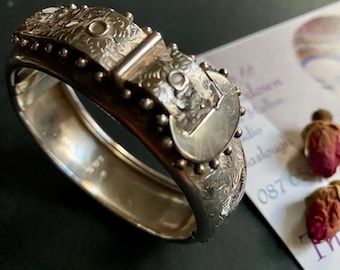 May include: A silver hinged bangle bracelet with intricate floral and geometric patterns. The bracelet features a buckle-style clasp and is adorned with small, raised circular details. The design is detailed and ornate, suggesting a vintage or antique style.