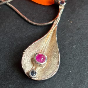 May include: A silver-toned pendant with a textured, teardrop-shaped surface featuring a central round pink gemstone and a smaller dark blue gemstone below it. The pendant is attached to a thin silver-toned chain, with a small blue stone at the bail.
