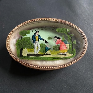 May include: An oval-shaped antique trinket box with a detailed miniature painting under glass. The painting depicts a man in a blue jacket and white pants, a woman in a red dress, and a small figure in a green coat. The box has a gold-colored beaded border.