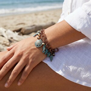 May include: A copper-toned charm bracelet featuring sea-themed charms. The bracelet includes charms shaped like seahorses, shells, and a starfish. The charms are in shades of turquoise and white, with a rustic, beachy aesthetic.