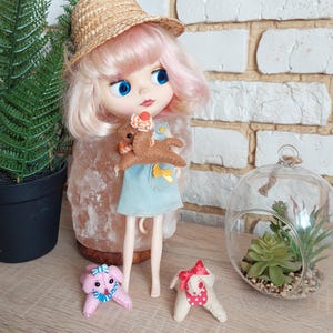 Puede incluir: Una muñeca con cabello rosado y ojos azules que lleva un sombrero de paja y un vestido azul, sosteniendo un perro de fieltro marrón. Dos pequeños perros de fieltro están sobre la mesa. También se ve un pequeño terrario con una planta suculenta.