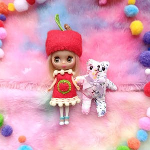 Puede incluir: Una muñeca con sombrero y vestido de manzana roja con diseño de fresa, junto a un pequeño osito de peluche con lazo. El fondo es una superficie colorida y esponjosa con un borde de pompones de colores.