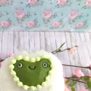 May include: A plush, white frog-shaped pillow with a green face, feet, and a heart-shaped border. The frog has large, round eyes and a smiling mouth. The pillow is decorated with a green bow.