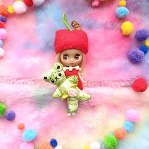 Puede incluir: Una muñeca con un sombrero de manzana roja y un vestido rojo, sosteniendo un juguete de dinosaurio verde y blanco. La muñeca tiene el pelo rubio y está rodeada de pompones de colores sobre un fondo esponjoso de arcoíris.