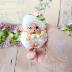 May include: A small, plush doll with a whimsical design. The doll features a white, fluffy body, a white hat with a pink rose, and a light green collar. It has a painted face with blue eyes and freckles. The doll's arms and feet are visible.