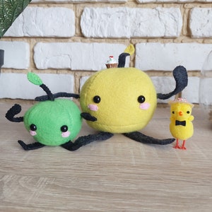 Puede incluir: Una creación de fieltro caprichosa con un personaje verde y otro amarillo con forma de fruta, con extremidades y rasgos faciales negros. Un pequeño pollito con pajarita y sombrero está cerca. Una pequeña magdalena está encima del personaje amarillo.