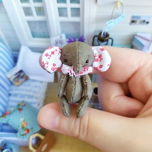 May include: A miniature stuffed elephant doll with gray fabric and patterned ears. The doll has a ruffled collar and is held in a person's hand. The background shows a miniature house with a window and decorative items.