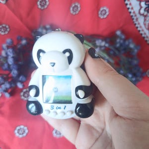 May include: A white and black panda-shaped digital handheld game console featuring a small square screen, four circular control buttons, and the text 3 in 1 printed in blue on the front. The device has a smooth plastic finish and a small metal loop.