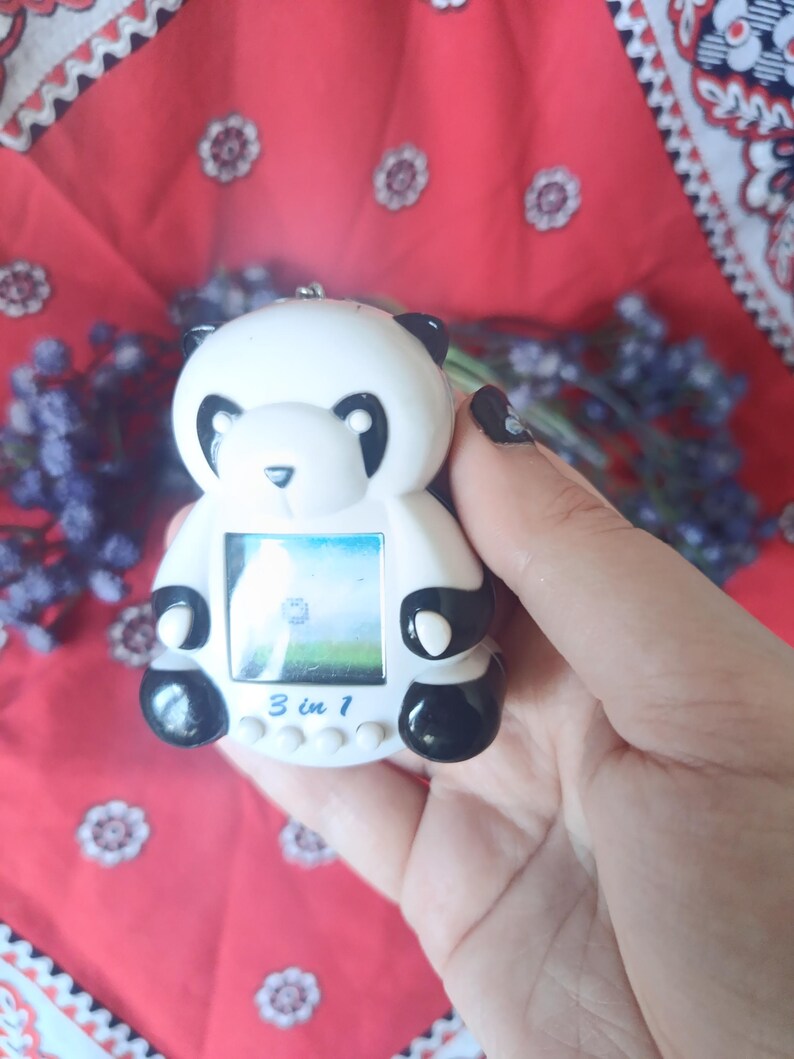 May include: A white and black panda-shaped digital handheld game console featuring a small square screen, four circular control buttons, and the text 3 in 1 printed in blue on the front. The device has a smooth plastic finish and a small metal loop.
