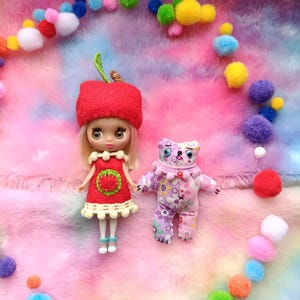 Puede incluir: Una muñeca con sombrero y vestido de manzana roja con diseño de manzana verde, junto a una muñeca de oso morado con estampados florales. Las muñecas están rodeadas de pompones coloridos sobre un fondo mullido multicolor.