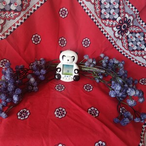 May include: A white and black panda-shaped digital pet toy with a small screen and buttons labelled 3 in 1. The device features a rounded head, black ears, and black eye patches, positioned centrally among sprigs of small, dried purple flowers.
