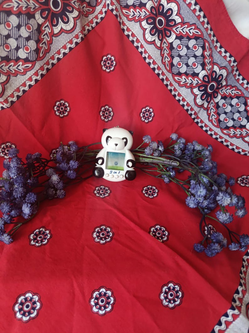 May include: A white and black panda-shaped digital pet toy with a small screen and buttons labelled 3 in 1. The device features a rounded head, black ears, and black eye patches, positioned centrally among sprigs of small, dried purple flowers.
