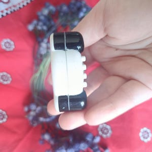 May include: A small, rectangular object with a white centre section and glossy black ends. The white side features a vertical row of four small, protruding circular buttons. The object has a smooth, rounded finish and a distinct seam separating the colours.