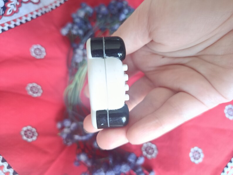 May include: A small, rectangular object with a white centre section and glossy black ends. The white side features a vertical row of four small, protruding circular buttons. The object has a smooth, rounded finish and a distinct seam separating the colours.