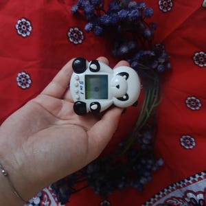 May include: A white and black panda-shaped digital pet toy with a small square screen and three circular buttons on the front. The device features a panda face design with black ears, eye patches, and a nose, with the text 3 in 1 printed on the front.