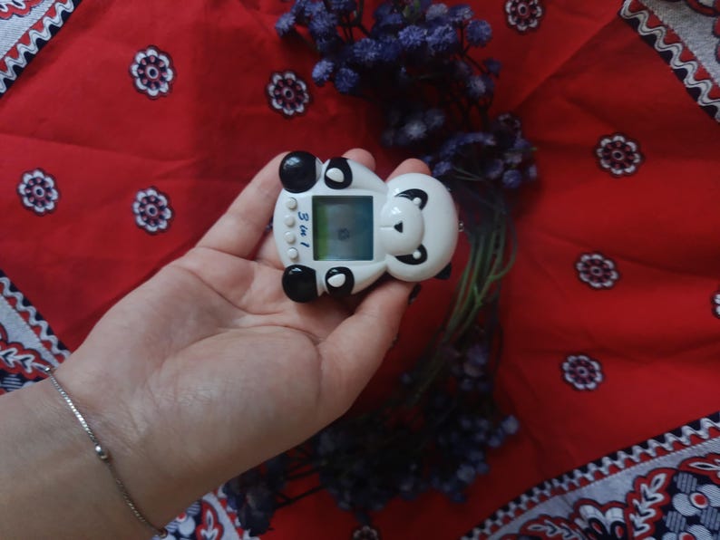 May include: A white and black panda-shaped digital pet toy with a small square screen and three circular buttons on the front. The device features a panda face design with black ears, eye patches, and a nose, with the text 3 in 1 printed on the front.