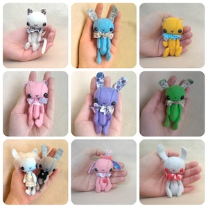 May include: A collection of nine small, handcrafted felt animal dolls in various colors including white, blue, yellow, pink, purple, and green. Each doll features large black button eyes, stitched facial details, and decorative fabric collars or accessories.