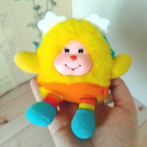 May include: A plush toy with a yellow, fluffy head, yellow arms, and blue feet. The toy has a smiling face with a red nose and a rainbow-colored belly. White cloud-shaped accents are on the head.