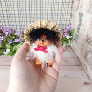 Könnte beinhalten: Eine kleine Puppe mit pfirsichfarbenem Gesicht, blauen Augen und einem kleinen roten Mund. Sie trägt einen strukturierten beigen Strohhut, schwarzes Kunstfellhaar und einen weißen Kunstfellkragen, der mit einer kleinen roten Schleife verziert ist.