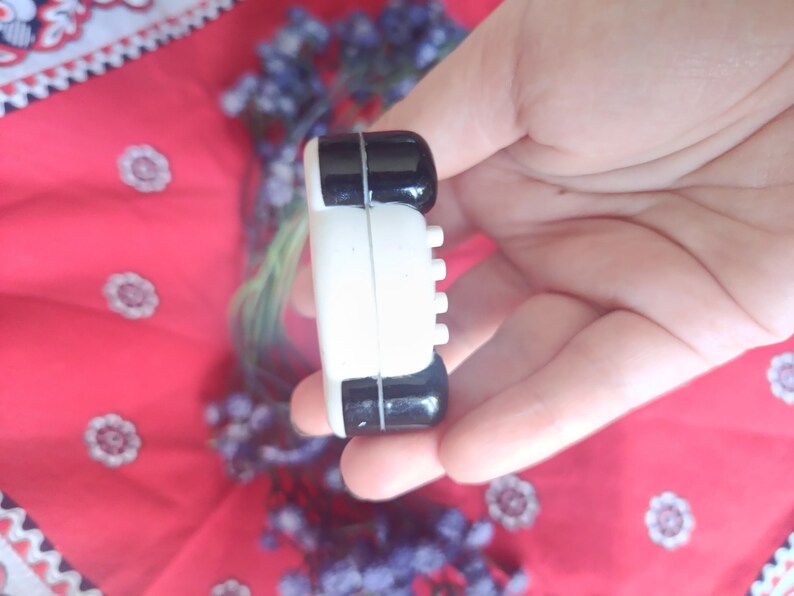 May include: A small, rectangular, two-toned object featuring a glossy black top and bottom section separated by a white centre band. The white section displays a series of four small, evenly spaced, protruding white circular buttons on the side.