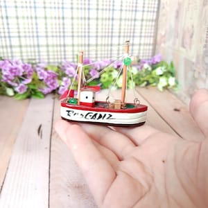 Puede incluir: Un pequeño barco de madera pintado de rojo, blanco y marrón, con el texto "Rio Cadiz" en el lateral. El barco tiene dos mástiles, una pequeña cabina y detalles verdes y rojos. Se sostiene en la mano de una persona.