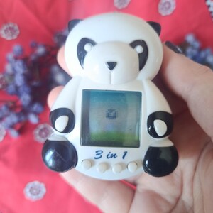 May include: A white and black panda-shaped digital handheld game console featuring a square screen displaying a pixelated graphic. The device has three small white circular buttons below the screen and the text 3 in 1 printed in blue.